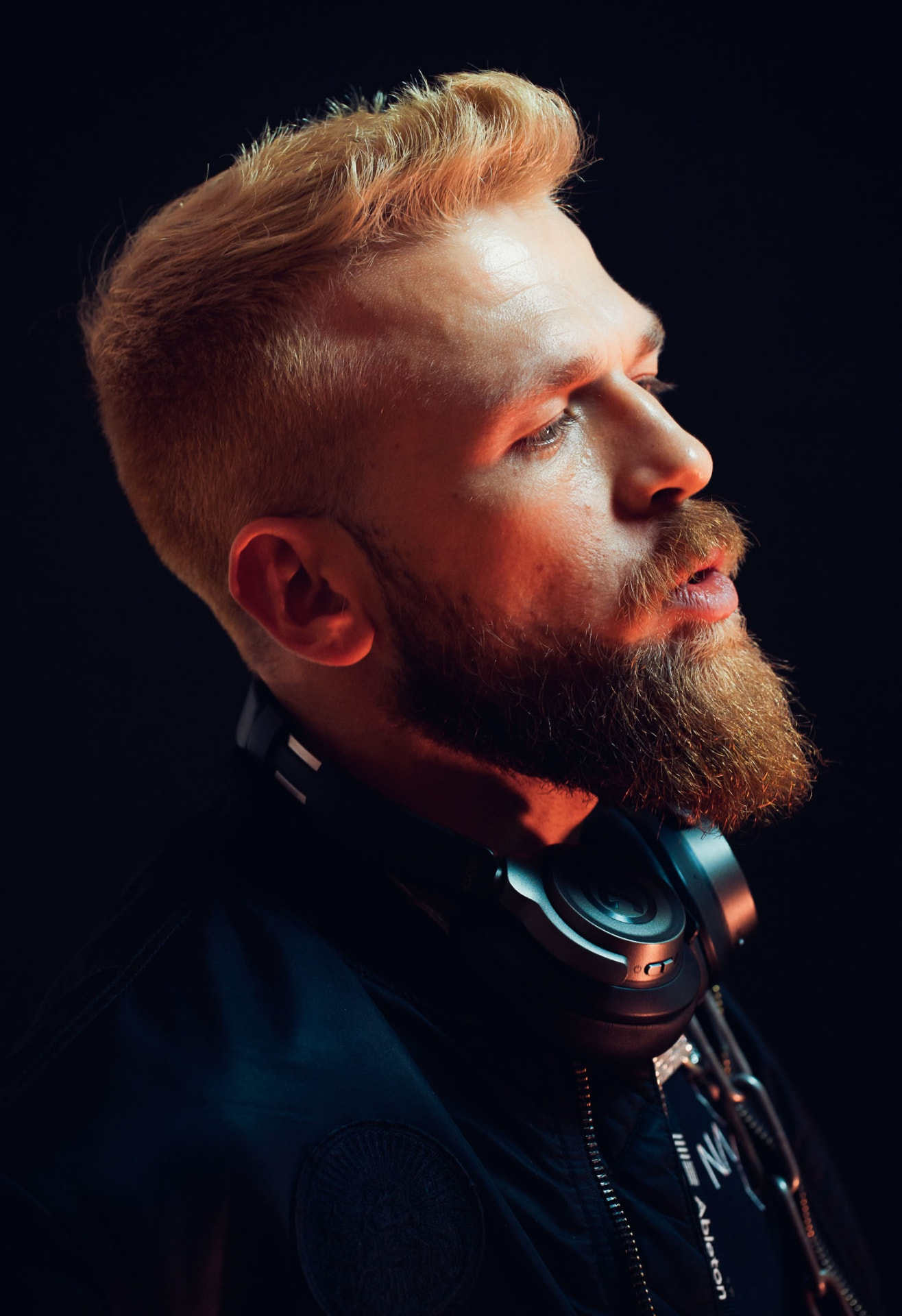 Backlit profile portrait of a bearded DJ with headphones around the neck
